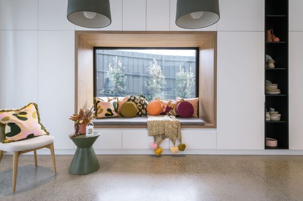 A bright contemporary built-in window-seat nook with a deep timber-lined reveal, colourful patterned and round cushions, and white integrated cabinetry. A terrazzo-style floor, sculptural green side table, pendant lights, and a tall dark open shelving unit complete the playful modern interior.