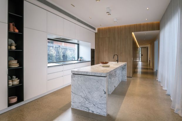 A refined contemporary kitchen with a large white-veined stone island, integrated white cabinetry, black open shelving, timber wall panelling, and a long circulation corridor. Full-height sheer curtains line the right side, while recessed and concealed lighting creates a warm, polished atmosphere.