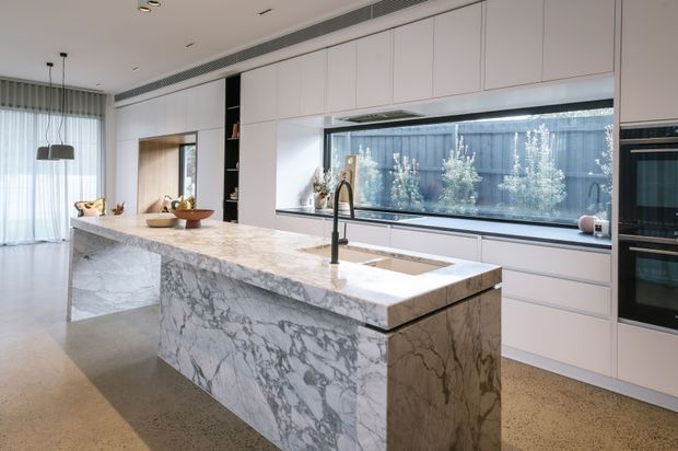 A bright contemporary kitchen with a large white marble waterfall island, integrated sink and tall black tap. Full-height pale cabinetry surrounds built-in appliances, with a long horizontal window above the rear benchtop looking onto a planted courtyard. The open-plan space includes sheer curtains and dining pendant lights, with polished concrete or terrazzo-like flooring and restrained neutral finishes.