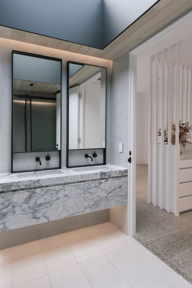 A refined contemporary bathroom featuring a floating double vanity with a dramatic white-and-grey veined stone benchtop and fascia, black wall-mounted taps, and two tall black-framed mirrors. Pale tiled flooring, light grey wall surfaces, warm timber ceiling lining, and a dark ceiling create a high-contrast, elegant fit-out. A white fluted screen and open doorway reveal an adjacent hallway or room.