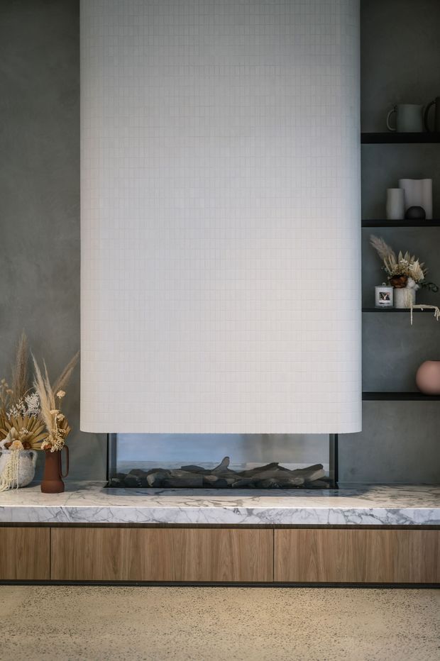 A refined contemporary living-room fireplace feature with a large curved white tiled chimney hood or surround, dark grey wall finish, open shelving with decorative objects, a marble hearth or benchtop, and warm timber cabinetry below. A linear firebox with dark stones sits beneath the curved feature, while a terrazzo-like floor grounds the composition.