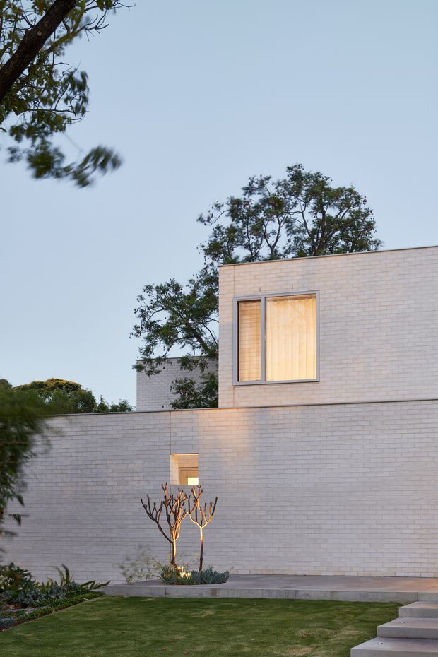 Exterior architectural photograph of a minimalist contemporary residence at dusk. The building has clean rectilinear volumes, a flat roof, pale white brick walls, dark-framed windows with warm interior light, and a simple landscaped courtyard with lawn, raised planting beds, and steps.