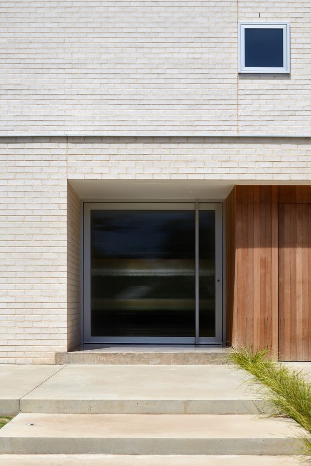 A close-up exterior view of a contemporary residential entrance. The facade is clad in pale horizontal brick, with a recessed entry containing a dark glazed aluminium-framed door, a small high-set window, and a warm vertical timber-clad panel beside the doorway. Concrete steps and paving lead to the entrance, with ornamental grass at the edge.