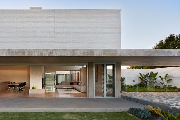 Exterior view of a contemporary flat-roof home with a clean white brick upper volume, broad concrete canopy, full-height glazed openings, and an open ground-level living and dining area. A paved courtyard with tropical planting and glass balustrade is visible to the right.