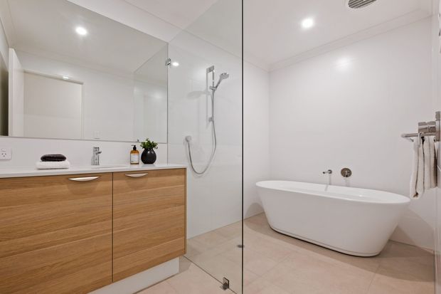 Bright contemporary bathroom with a freestanding white bathtub, frameless glass shower enclosure, wall-mounted timber vanity with white benchtop, large mirror, and pale tiled flooring and walls. The room has recessed ceiling lighting and a clean, minimal fit-out.