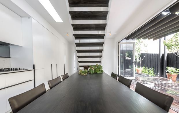 Bright contemporary open-plan dining and kitchen space with a long dark timber dining table, matching upholstered chairs, streamlined white kitchen cabinetry, and a dramatic open-riser stairway overhead. Full-height glazing with dark roller blinds opens to a timber deck and planted courtyard.