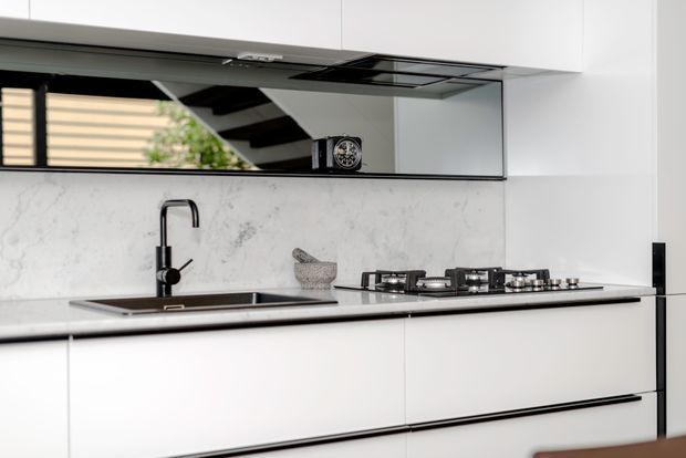 A sleek contemporary kitchen with handleless white cabinetry, a black stone or composite benchtop, black undermount sink and tap, gas cooktop, and a white marble-look splashback. A wide mirrored splashback reflects the room and staircase, adding depth. The space has a clean, minimalist monochrome palette with subtle grey veining and black accents.