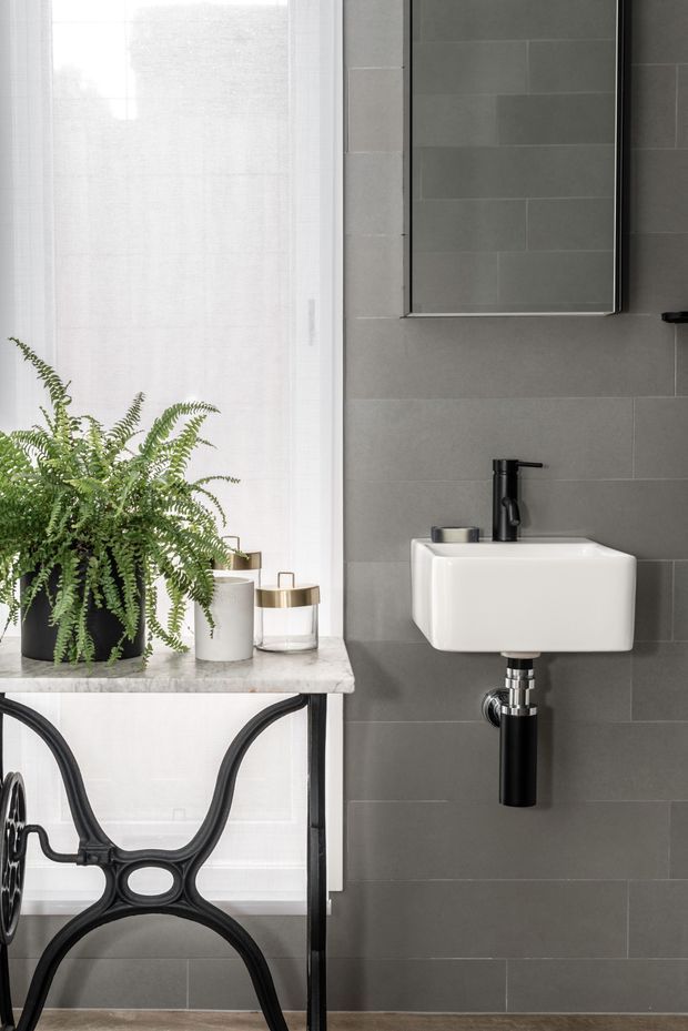 A compact, contemporary bathroom with a wall-mounted white basin and matte black tap and exposed waste pipe. Large grey rectangular wall tiles create a restrained backdrop, while a black-framed mirror hangs above. A marble-topped black metal console with a fern and decorative containers sits beside a bright window with sheer curtains. A partial black sculptural stool or chair is visible in the foreground.