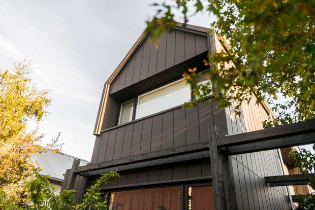 Exterior view of a contemporary multi-storey residence with a steep gabled roof, dark vertical cladding, large horizontal glazing, and a timber-framed lower-level structure. Mature trees partially screen the facade.