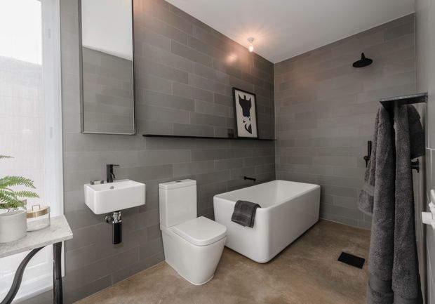 Contemporary grey-tiled bathroom with a wall-mounted white basin, toilet, freestanding bathtub, black fixtures and shelf, and a walk-in shower area. A large window, mirror, towel storage, and potted plant add brightness to the compact space.