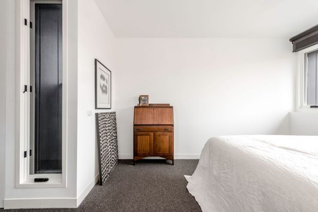 Bright, compact bedroom with white walls, a white upholstered or linen-covered bed, grey carpet, and a warm mid-century-style wooden secretary cabinet. Tall narrow glazing and a small window provide natural light, while framed artwork and a patterned panel add restrained decorative detail.