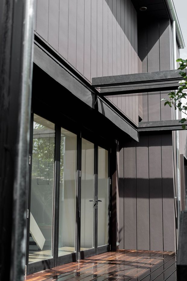 A close-up exterior view of a contemporary multi-level building or entry courtyard. Dark vertical-panel cladding, black-framed full-height glazing and doors, projecting balcony or canopy structures, exposed metal drainage and a wet tiled or brick-paved surface create a sharply detailed modern facade.