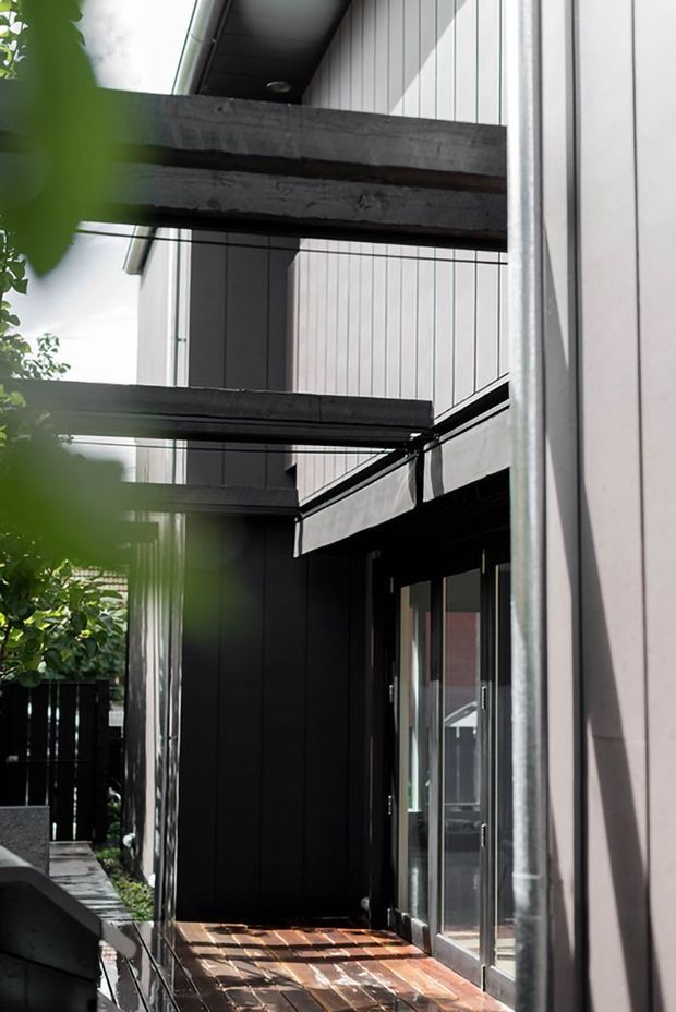 A narrow, contemporary exterior passage or deck beside a vertically clad building. Dark timber or metal beams project overhead, with black-framed glazed doors, timber decking, and contrasting light vertical wall cladding. Dense greenery partially frames the view.