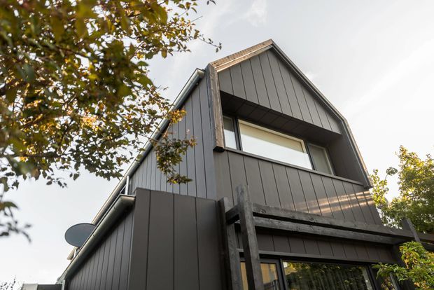 Low-angle exterior view of a contemporary multi-storey house with a steep gabled roof and dark vertical cladding. The facade includes a recessed upper-level window, exposed timber-like structural framing, and lower-level glazing, partially screened by mature trees.