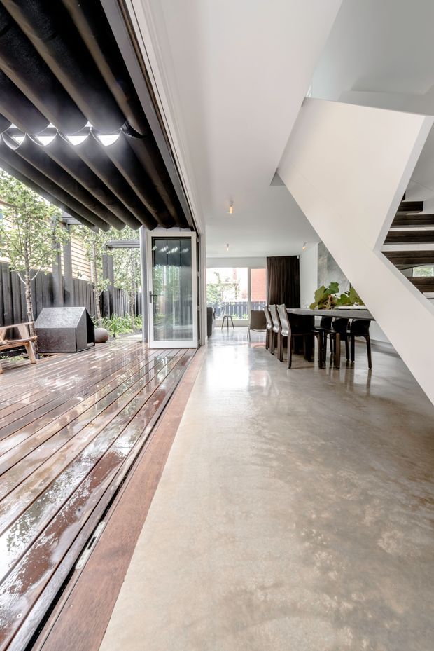 A contemporary open-plan interior connects directly to a timber outdoor deck through large glazed sliding doors. The space includes a dining area beneath a prominent white geometric staircase, polished concrete flooring, white walls and ceiling, dark timber or metal stair treads, and a covered outdoor patio with exposed dark beams. The image has a bright, refined indoor–outdoor character.