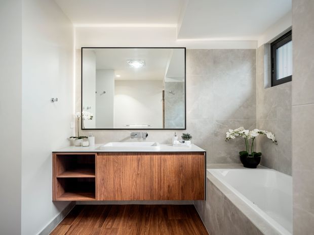 Bright contemporary bathroom with a floating timber vanity, integrated white basin, large frameless mirror, freestanding-style bathtub, and light stone-look wall tiles. A narrow black-framed window provides natural light, while recessed and concealed lighting create a clean, refined interior.