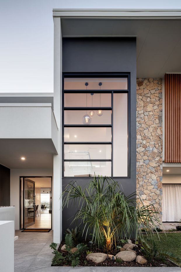 Exterior architectural photograph of a contemporary multi-level residence, showing a tall black-framed window, dark rendered walls, pale stone feature cladding, vertical timber screening, and a landscaped entry garden with tropical planting and rocks. A glazed entry door reveals a dining or living area inside.