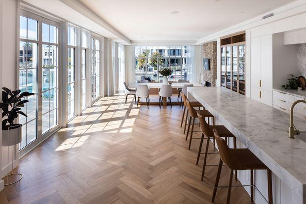 Bright, spacious open-plan kitchen and dining area with floor-to-ceiling glazing, a long marble island with timber bar stools, white built-in cabinetry, and warm herringbone timber flooring. The room overlooks a waterfront or marina setting through the windows and has refined contemporary detailing.