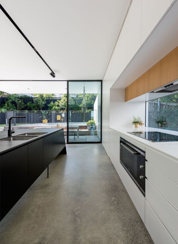 A bright, contemporary galley kitchen with a black island, integrated sink and tap, white full-height cabinetry, warm timber overhead cupboards, and a white stone benchtop. The polished concrete floor and ceiling-mounted black linear track light add an industrial-modern touch. Full-height glazing and a glass door open to a landscaped outdoor area and patio beyond.