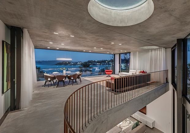 Contemporary open-plan upper-level living and dining space overlooking a waterfront setting. The room features a raw concrete ceiling and floor, expansive floor-to-ceiling glazing, sheer curtains, a sculptural curved stair with slim metal balustrade, and a large circular skylight or ceiling feature. A dining table and lounge seating open onto a terrace with water views.