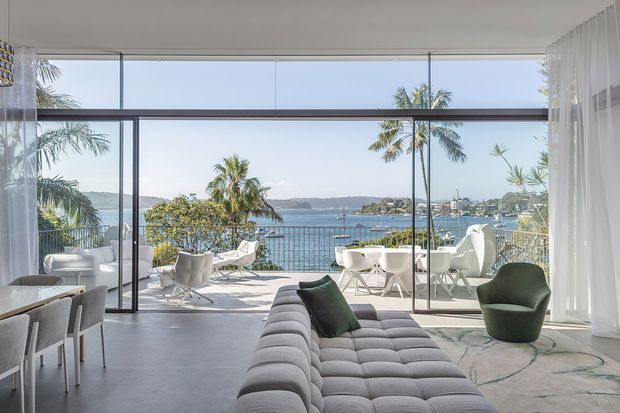 Bright, contemporary open-plan living space with floor-to-ceiling glazing overlooking a waterfront setting. A large tufted sectional sofa faces the view, with sculptural lounge chairs, a dining area, sheer curtains, and a spacious terrace beyond the fully glazed opening. The room has a refined, airy coastal feel with strong indoor-outdoor connection.