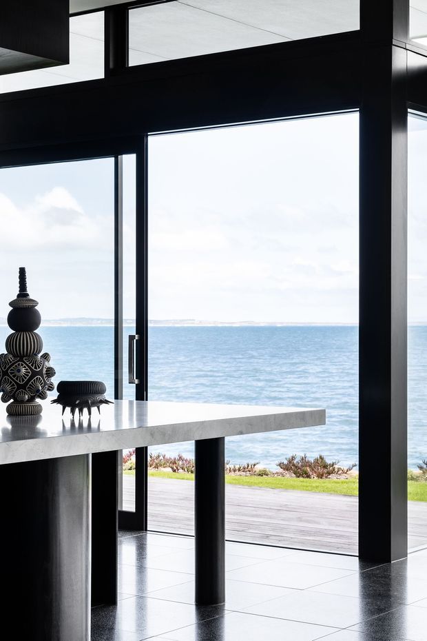 A dramatic contemporary interior opening directly onto a waterfront, with floor-to-ceiling glazing, black structural framing, a polished dark tiled floor, and a substantial pale stone island or counter. Sculptural black decorative objects sit on the counter, while a timber deck and ocean horizon are visible beyond.
