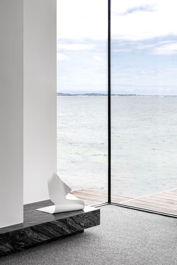 Minimalist interior corner opening directly onto a waterfront through floor-to-ceiling glazing. White walls surround a dramatic dark veined stone slab ledge or bench, with a sculptural white chair and grey carpet. A timber deck extends outside over calm water, creating a bright, serene, highly pared-back composition.