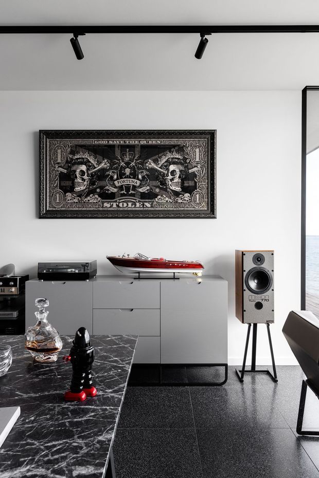A sophisticated contemporary interior vignette featuring a monochrome media and dining area. A framed black-and-white artwork hangs above a streamlined light-grey sideboard displaying a turntable and a decorative red model boat. A vintage-style speaker stands beside it, while a black marble dining table with a glass decanter and sculptural ornament occupies the foreground. Black track lighting, dark flooring, and expansive glazing reinforce the refined, high-contrast aesthetic.