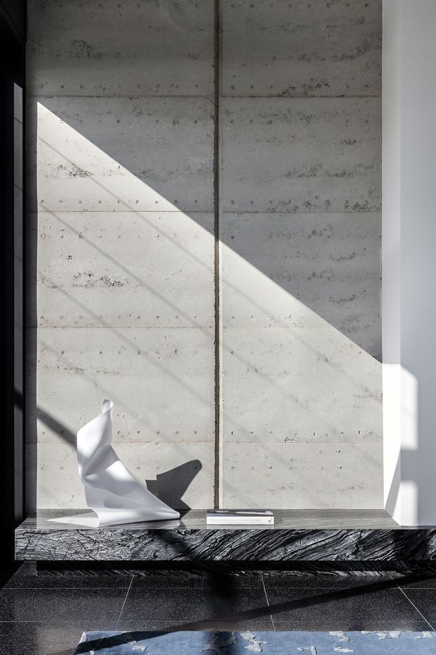 A minimalist interior detail featuring a large concrete panelled wall illuminated by strong geometric sunlight and shadow. A dark, dramatically veined stone ledge runs along the base, displaying a sculptural white object and a small book or box. The floor is dark tiled, with part of a blue patterned rug visible in the foreground. The image has a refined, architectural and high-contrast character.