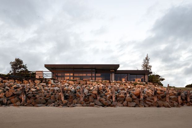 Low, single-storey contemporary house viewed from the exterior, set behind a substantial rocky retaining wall. The building has dark horizontal cladding or framing, extensive glazing, and a broad projecting flat roof beneath an overcast sky.