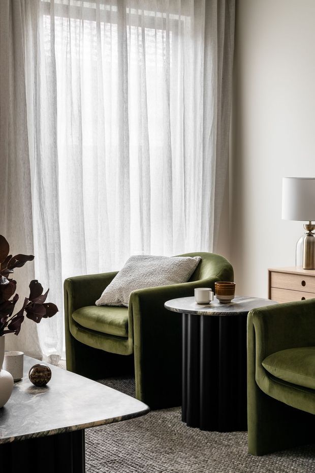 A refined living room corner with tall sheer curtains, two deep olive-green upholstered armchairs, a black fluted cylindrical side table, and a pale stone coffee table. Neutral walls, a textured grey rug, sculptural lamp, and understated decor create a soft, contemporary atmosphere with natural filtered light.