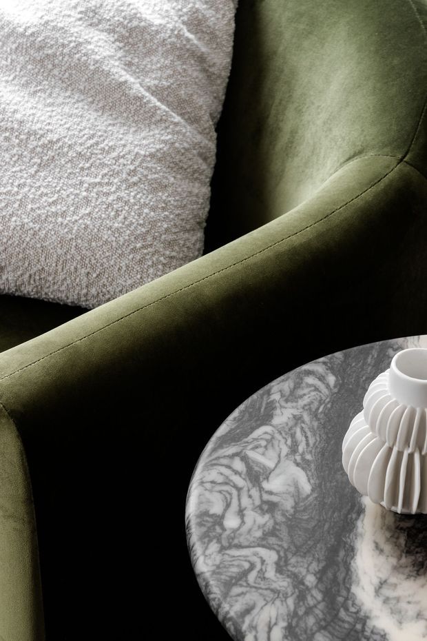 Close-up interior vignette featuring a deep olive-green upholstered armchair with visible stitching, a textured pale grey cushion, and a round grey-and-white veined stone or marble side table holding a sculptural white ceramic vase. The composition is intimate, refined, and softly lit, emphasizing rich textures and organic veining.