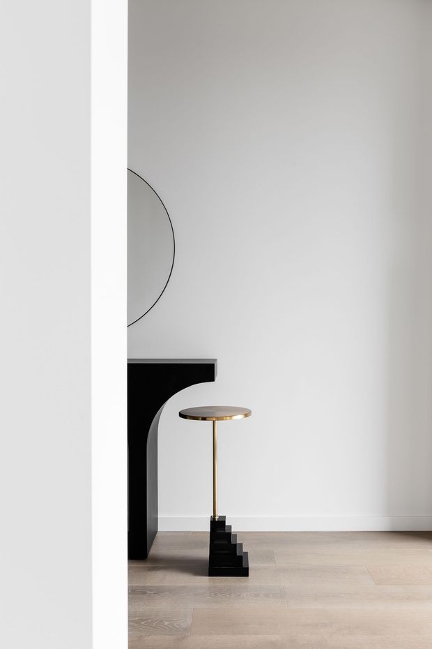 Minimalist interior vignette featuring a pale timber floor, white walls and a sculptural black console or fireplace-like architectural feature with a curved support. A slender brass-and-black pedestal lamp stands beside it, while a partial oversized circular wall artwork or mirror is visible. The composition is sparse, monochromatic and refined.