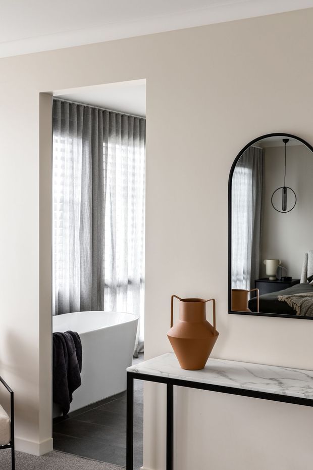 A refined interior view showing a marble-topped console table with black metal framing, a terracotta decorative vase, and a large arched black-framed mirror. Through the wide opening is a bathroom with a freestanding white bathtub, dark floor tiles, and layered grey sheer curtains. The neutral beige walls, white ceiling trim, and restrained furnishings create a calm, contemporary setting.