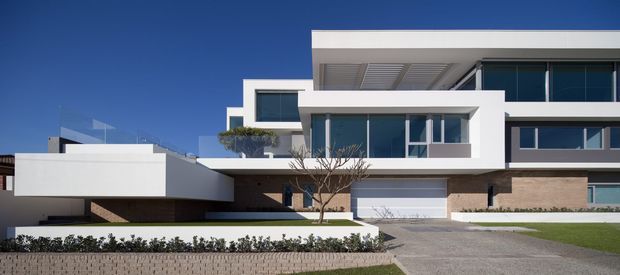 Street-facing exterior view of a large multi-level contemporary residence with crisp white cubic volumes, extensive dark-framed glazing, recessed balconies, a flat roof, and a broad driveway leading to an integrated garage. Low landscaped planting and a rendered masonry retaining wall define the foreground.