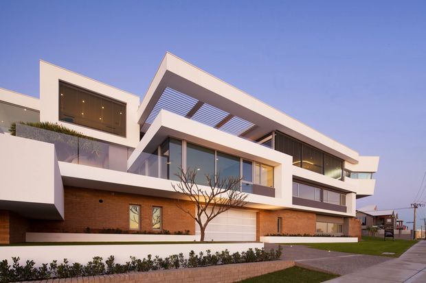 Exterior photograph of a large contemporary multi-level residence with strong geometric volumes, projecting balconies, extensive glazing, a covered pergola, white rendered surfaces, and a warm brick base. The landscaped front garden, driveway, garage, and suburban streetscape are visible.