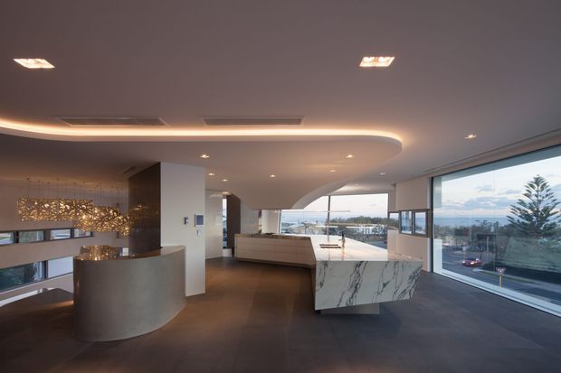 A sophisticated open-plan kitchen and living area with sweeping curved ceiling details, dark flooring, large marble-look island, curved cabinetry, sculptural pendant lighting, and expansive floor-to-ceiling glazing overlooking a coastal or urban landscape.