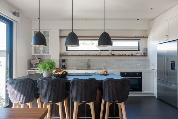 A bright contemporary kitchen with a large island and four dark woven bar stools. The room features white framed cabinetry, a pale stone-look splashback, dark flooring, stainless-steel appliances, open timber shelving, and three black pendant lights. A window and glazed door provide natural light.
