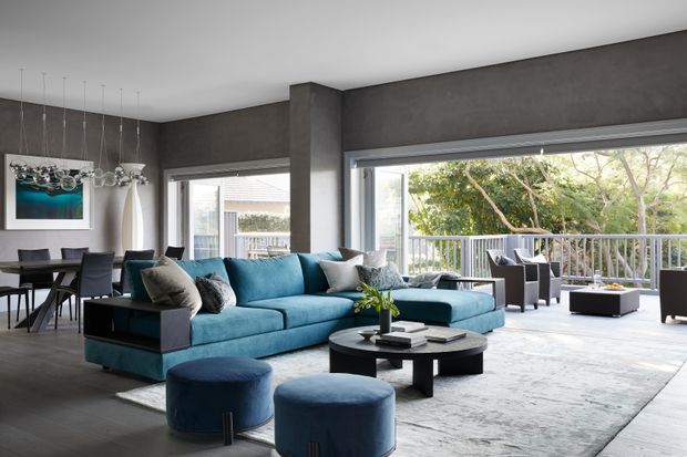 A spacious contemporary open-plan living and dining area with a deep teal sectional sofa, two rounded ottomans, a black circular coffee table, and a large pale rug. Dark textured grey walls, broad aluminium-framed windows and sliding doors, and a covered outdoor deck visible beyond create a modern indoor-outdoor connection. A dining table and sculptural pendant lighting occupy the left side of the room.