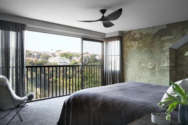A contemporary bedroom with a large bed, dark grey bedding and carpet, expansive sliding glass doors opening to a balcony, sheer and dark curtains, and a black ceiling fan. A botanical-patterned feature wall adds a textured, nature-inspired accent, while the balcony overlooks a leafy suburban landscape.