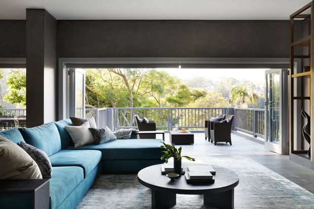 Contemporary living room with a large teal sectional sofa, round black coffee table, dark feature wall, open shelving, and wide glazed doors opening onto a balcony or terrace. The room is bright and spacious, with abundant greenery visible outside and a relaxed indoor-outdoor connection.