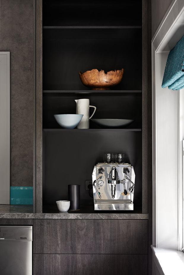 Close-up of a built-in coffee station within dark timber cabinetry. Open black shelving displays a wooden bowl, ceramic cups and bowls, wine glasses, and a stainless-steel espresso machine, with an integrated appliance partially visible below. The cabinetry is framed by dark grey stone-look surfaces and positioned beside a bright window.