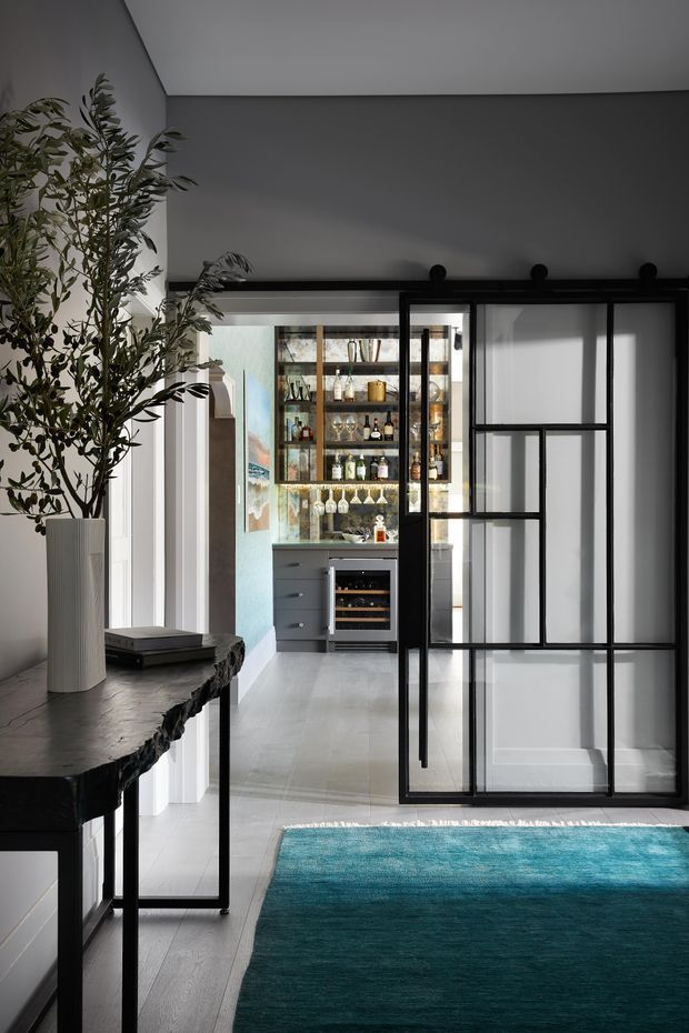 Interior view looking through black-framed glazed sliding doors toward a compact built-in home bar. The space has dark grey walls and ceiling, pale timber or light-toned flooring, black metal-framed glazing and joinery, a sculptural stone-topped console table with a tall indoor plant, and a large teal rug. The bar includes illuminated glass shelving, bottles and glassware, lower drawers, and a small wine refrigerator. The overall look is refined, moody, and contemporary with strong industrial-style metal framing.