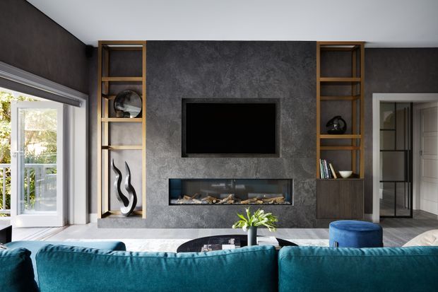 Contemporary living room with a dark textured feature wall incorporating a wall-mounted television and linear fireplace. Warm timber open shelving and dark cabinetry frame the feature wall, while a deep teal sectional sofa, blue ottoman, and large windows create a refined, dramatic interior.