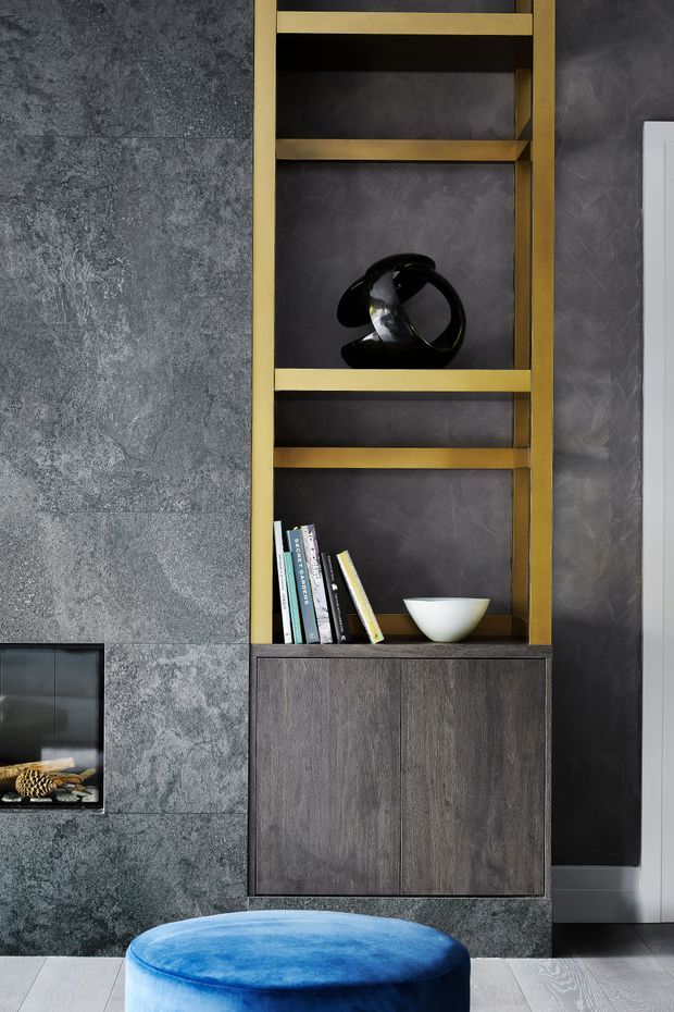 A close-up interior detail of a dark, sophisticated built-in shelving unit beside a fireplace. The shelving has bronze-gold metal framing, dark timber cabinetry, charcoal textured wall finishes, books, and sculptural decor. A vivid blue upholstered ottoman sits in the foreground, creating a strong accent against the moody grey and brown palette.