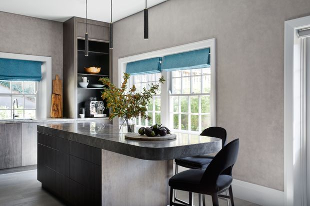 Contemporary kitchen with a dark timber island, rounded dark stone benchtop, integrated open shelving, grey textured walls, white-framed windows, and teal roller blinds. Three black pendant lights hang above the island, which is paired with two dark upholstered bar chairs.