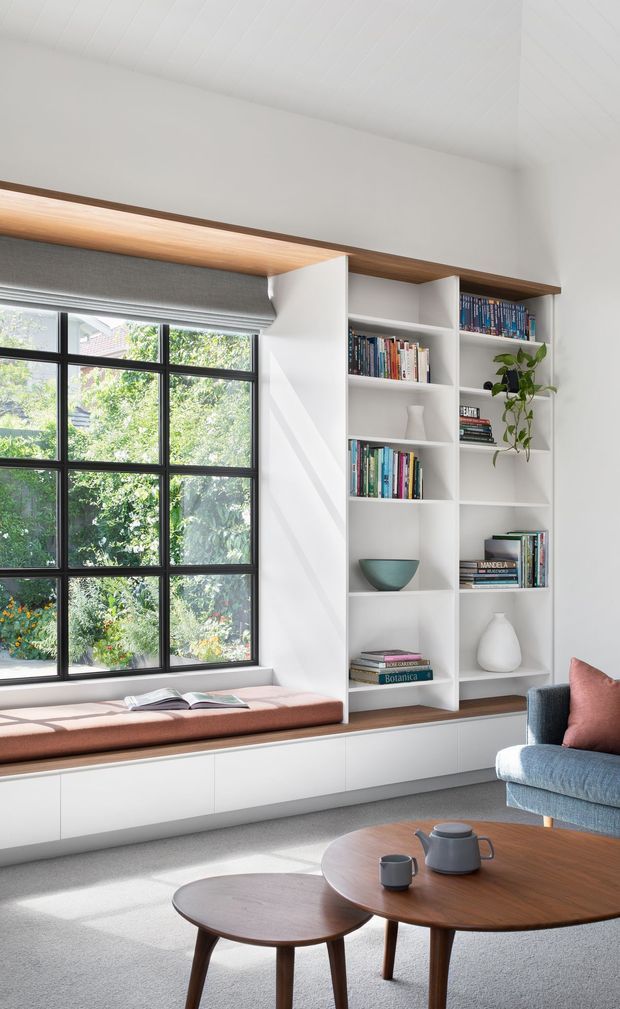 Bright, calm living room with a large black-framed grid window overlooking greenery, a built-in window seat with a muted terracotta cushion, and tall white open shelving integrated with warm timber cabinetry. The shelves display books, ceramics, and a trailing plant. A blue upholstered armchair and nested round timber coffee tables complete the space. The room has a white panelled ceiling, light grey carpet, and abundant natural light.