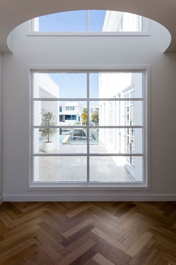 Bright interior view focused on a large white-framed multi-pane window overlooking a contemporary courtyard with white buildings and a pool or water feature. The room has white walls, a curved architectural opening above the window, and a dark-to-mid brown timber herringbone floor.