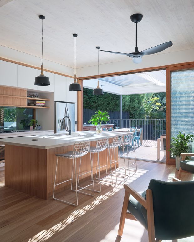Bright open-plan kitchen centred on a large timber island with a white benchtop and seating for several people. The space features white cabinetry, timber open shelving and joinery, black pendant lights, a black ceiling fan, and warm timber flooring. Large timber-framed glazing opens to a sunny covered outdoor terrace with garden planting and railings. Mid-century-inspired dining chairs and stools add a refined, warm character.