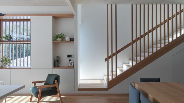 A bright, refined open-plan residential interior showing a timber staircase with slender vertical balusters, integrated white cabinetry and shelving, timber flooring, and a dining area in the foreground. A large glazed opening with vertical screening brings in natural light, while a mid-century style armchair and restrained greenery add warmth.
